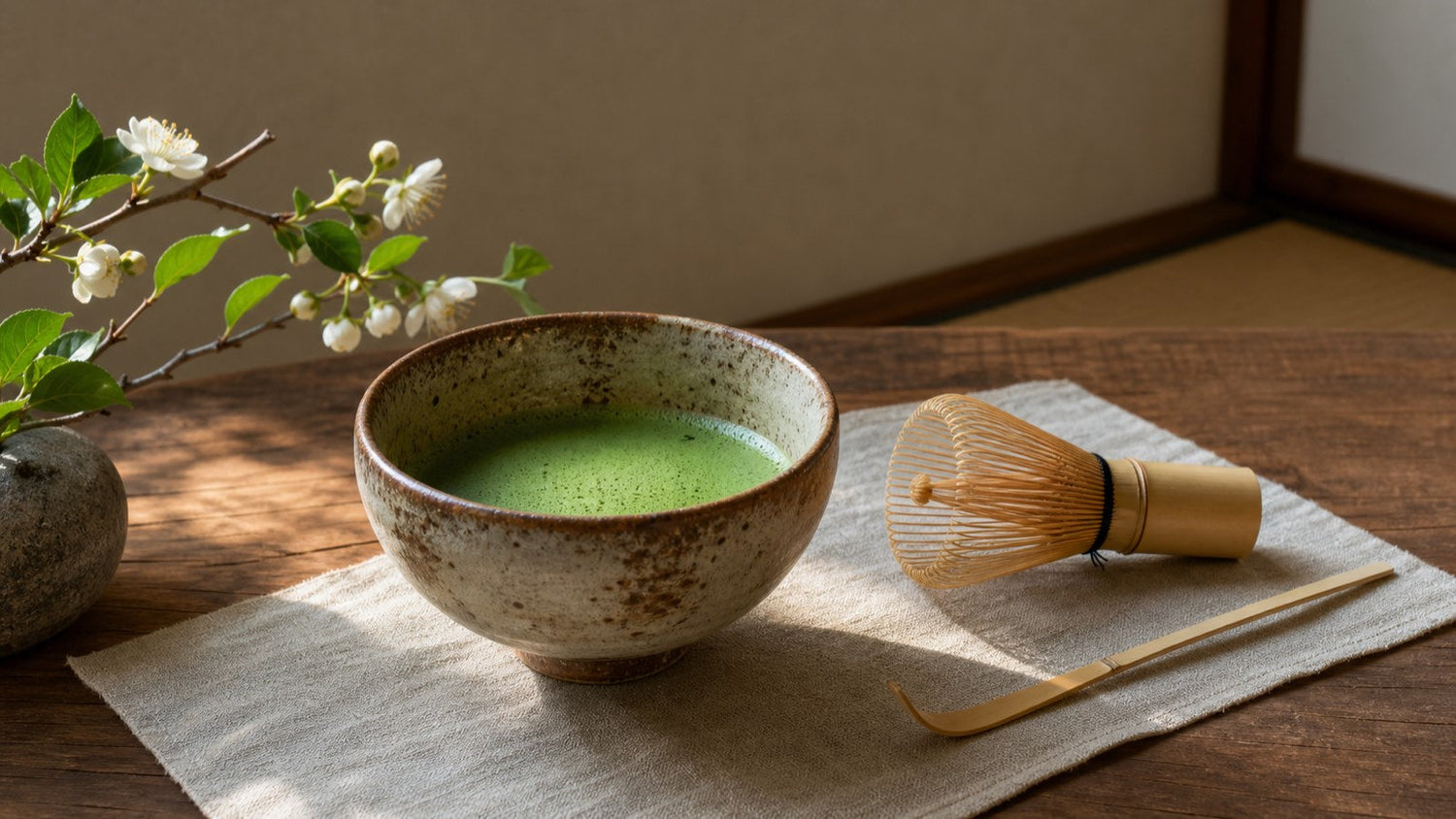 Ceramic bowl of whisked matcha with bamboo whisk, scoop and seasonal flower on a wooden tea table